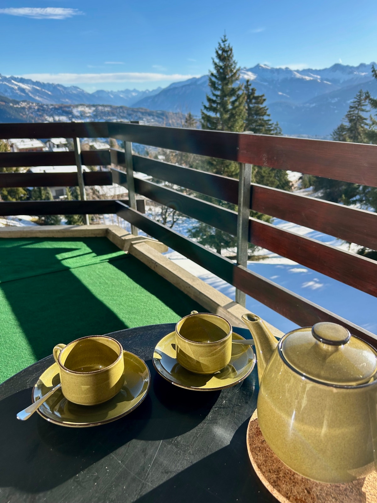 A cozy terrace setting features two green cups and a teapot arranged on a round table. A sweeping view of snow-covered mountains and trees is visible in the background, indicating a serene outdoor space. Bright sunlight enhances the scenic atmosphere.