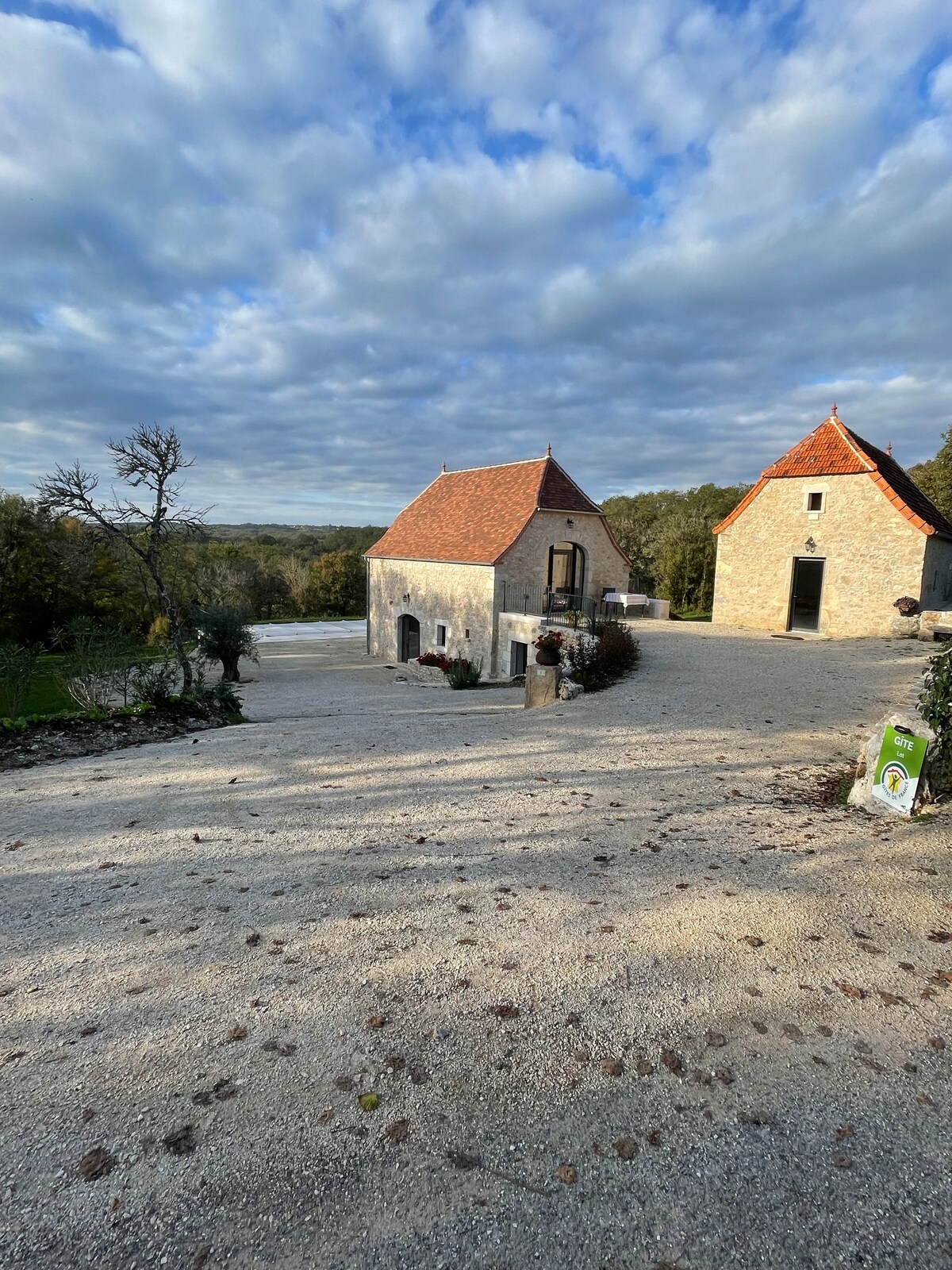 A charming outdoor view showcases two stone buildings with reddish-tiled roofs, surrounded by a gravel path. Lush trees are visible in the background, enhancing the serene countryside setting. The sky is partly cloudy, contributing to the tranquil atmosphere.