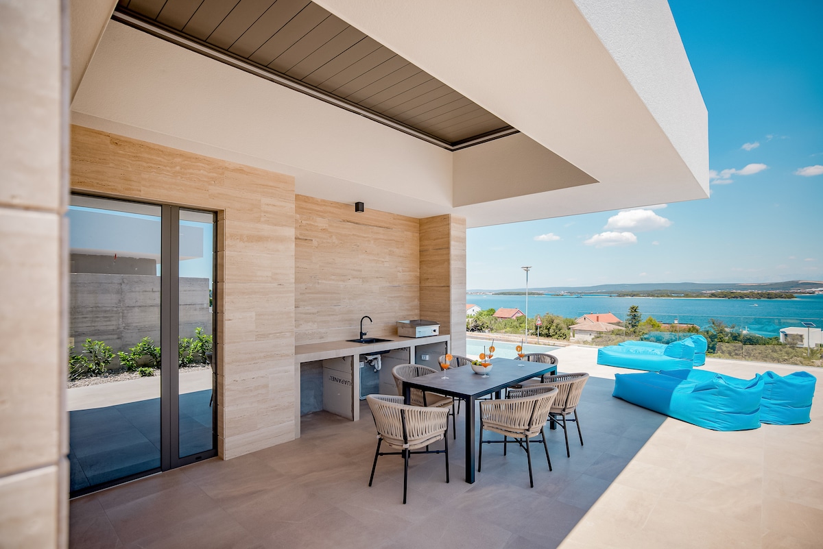 An outdoor terrace features a dining table surrounded by wicker chairs, with views of the sea in the background. An outdoor kitchen area is equipped with a sink and grill, while colorful bean bags are positioned for relaxation under a modern overhang.