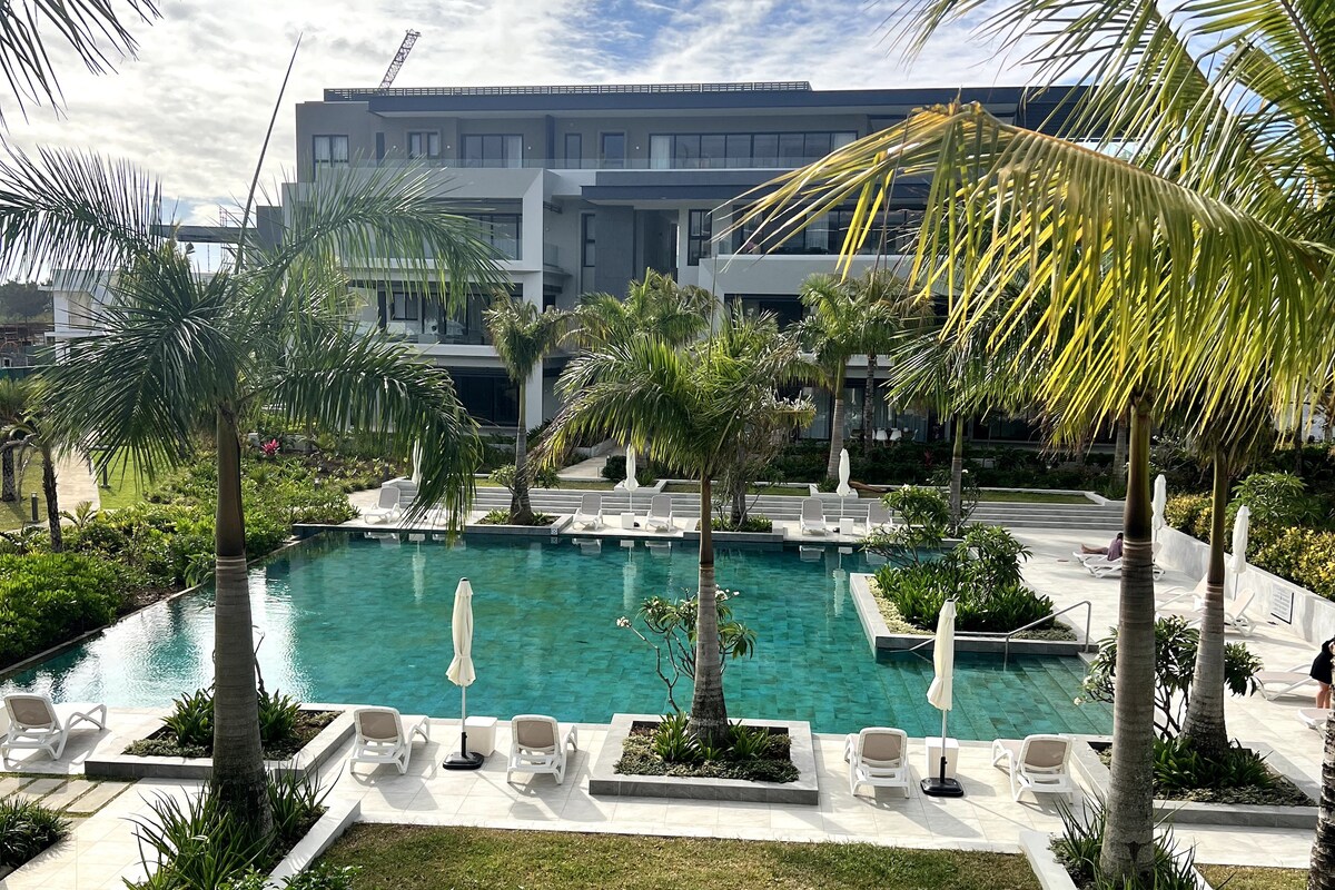A large swimming pool is surrounded by palm trees and tropical plants, with lounge chairs arranged along the edges. The backdrop features a modern building with multiple levels and large windows, under a partly cloudy sky.