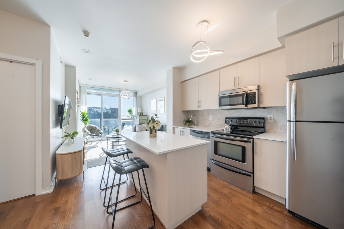 A bright kitchen and dining area features modern cabinetry and sleek stainless steel appliances. A central island with seating for four is complemented by natural light streaming through large windows, leading to a cozy lounge area with plants and comfortable furnishings.