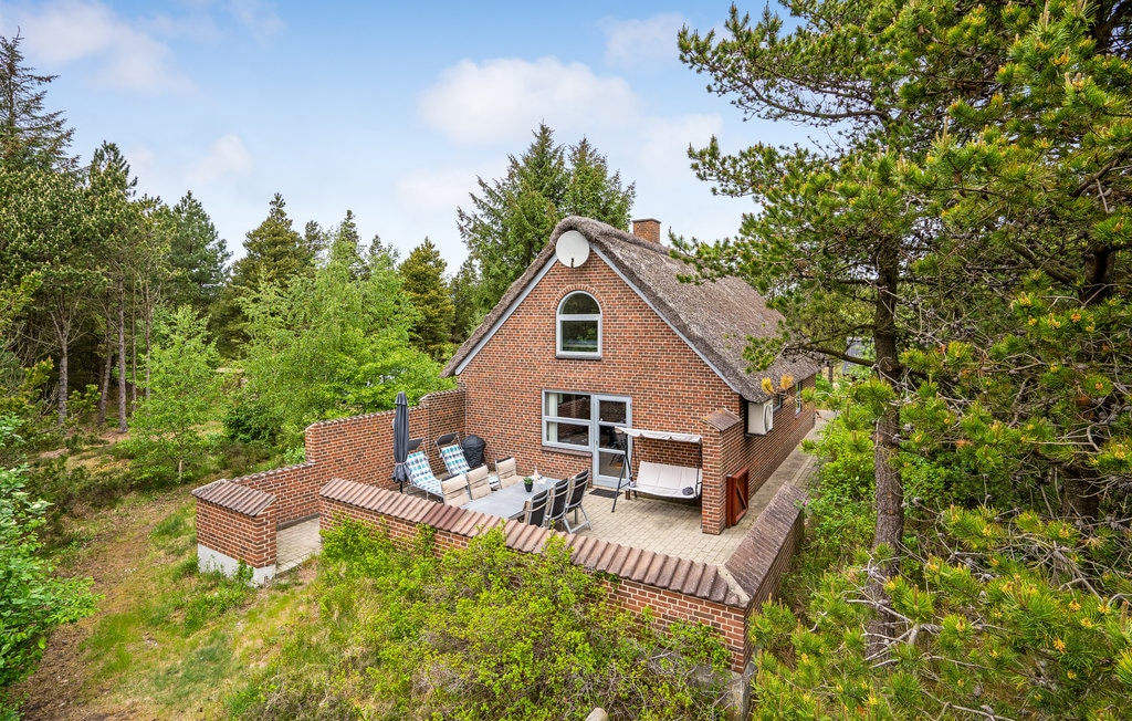 The exterior of a red brick house is surrounded by greenery, showcasing a spacious patio with multiple lounge chairs and tables. Tall trees stand nearby, offering a natural backdrop to the peaceful setting.