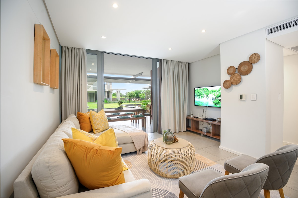 A inviting living area features a comfortable sectional sofa adorned with vibrant cushions. A flat-screen TV is mounted on the wall, and large glass doors provide views and access to the outdoor space. Natural light filters in through sheer curtains, enhancing the relaxed atmosphere.