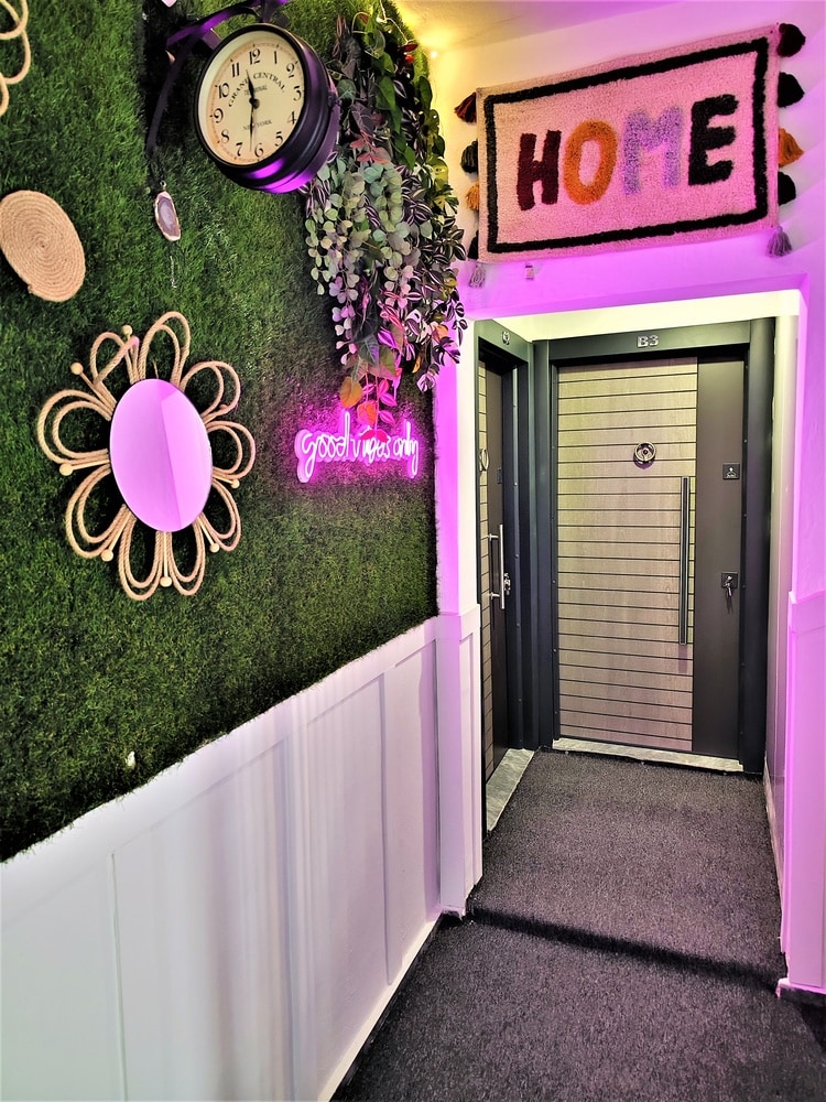 The entrance corridor features a decorative wall with greenery, a round mirror, and a clock. A soft carpet runs along the floor, leading to the elevator doors, which are framed by a colorful wall decor that includes the word 'HOME.'