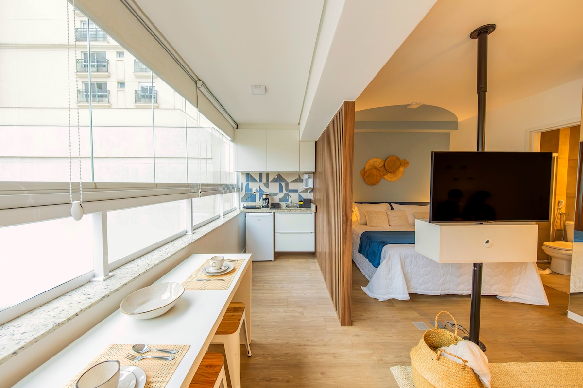 A spacious studio is shown featuring a modern design. Natural light enters through large windows, illuminating the open layout that includes a kitchen area and a sleeping space. A partition separates the sleeping area from the living space, enhancing privacy.