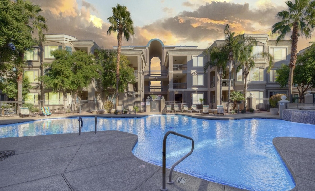 A large swimming pool is framed by palm trees and lush landscaping. Surrounding the pool are multiple seating areas and lounge chairs. The backdrop features a modern building with balconies overlooking the pool, creating a relaxed outdoor environment.