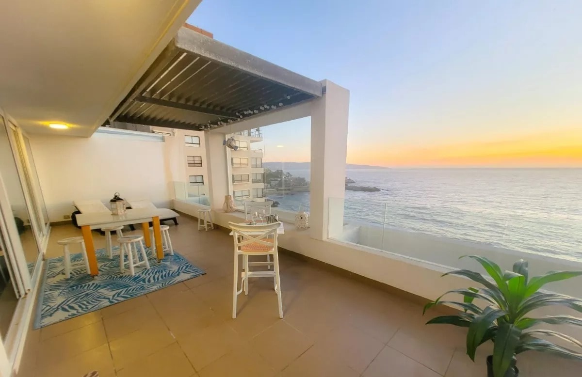 An outdoor terrace features a dining table surrounded by white chairs, overlooking the ocean at sunset. A cozy seating area with a light-colored futon is positioned under a shaded pergola. Lush greenery is present in a corner, contributing to a relaxing atmosphere.