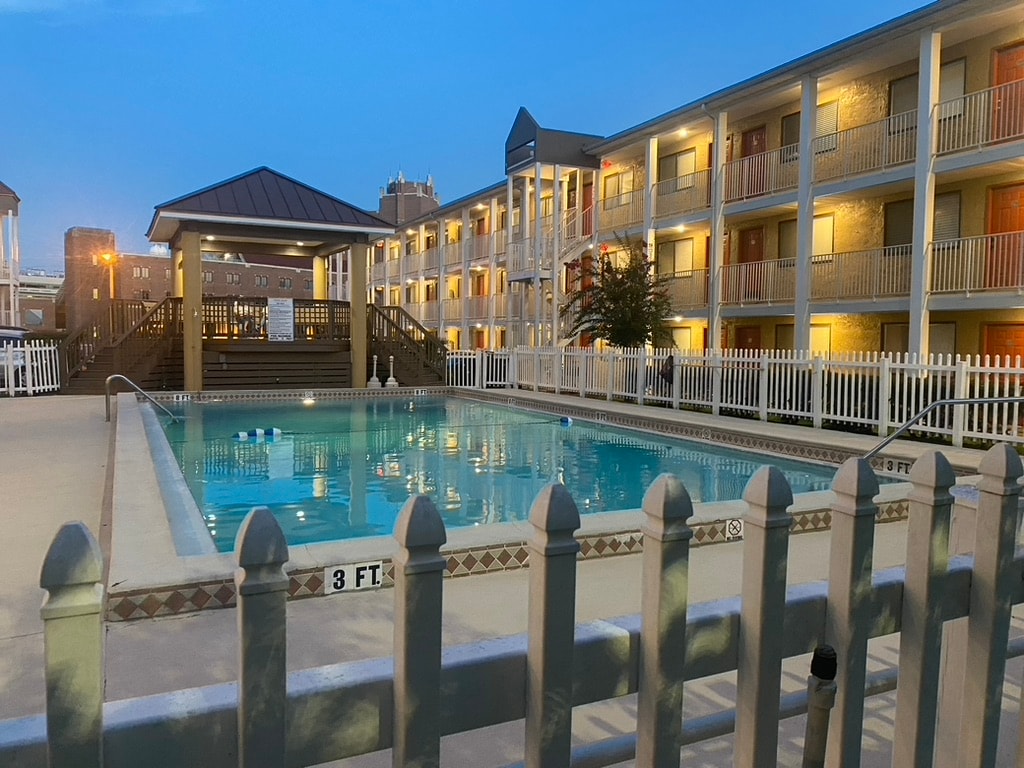An outdoor pool area is enclosed by a white picket fence, featuring a wooden gazebo at one end. The pool is inviting, with a shallow end marked at three feet. Surrounding the pool are two stories of accommodations, softly illuminated in the evening light.