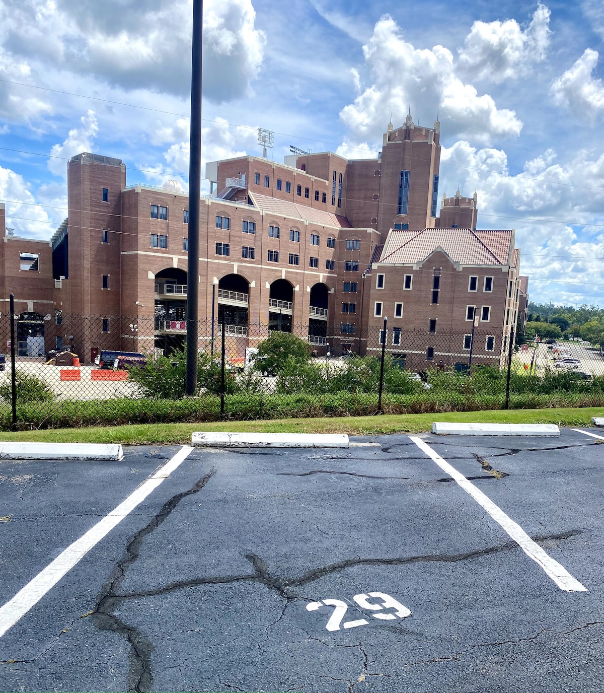 The image features a parking lot with a visible numbered space labeled '29'. In the background, a large, multi-story brick building is seen, adorned with distinct architectural elements. A chain-link fence partially encloses the area, while a blue sky dotted with clouds adds depth to the scene.