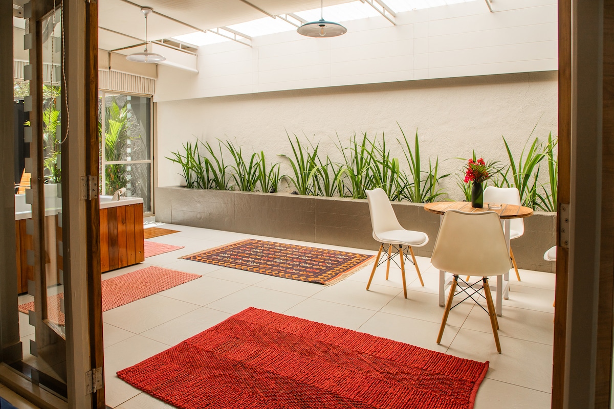 A bright outdoor patio area features white chairs surrounding a round table, complemented by vibrant red rugs. Lush green plants line the walls, and natural light is filtered through a skylight, creating an inviting space for relaxation.