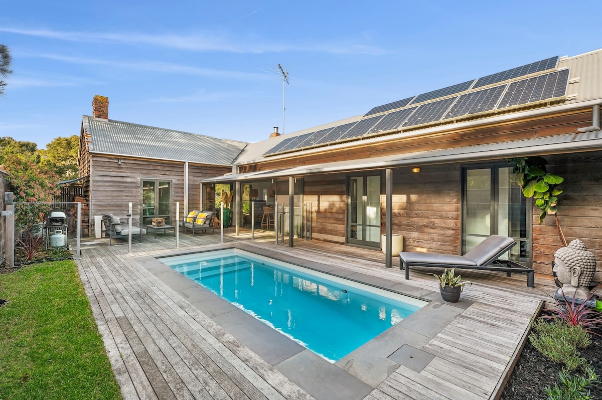 A modern outdoor area features a private solar heated pool surrounded by wooden decking. Comfortable loungers are positioned near the water, while lush greenery adds a touch of nature. Large glass doors provide access to the interior, blending indoor and outdoor living.