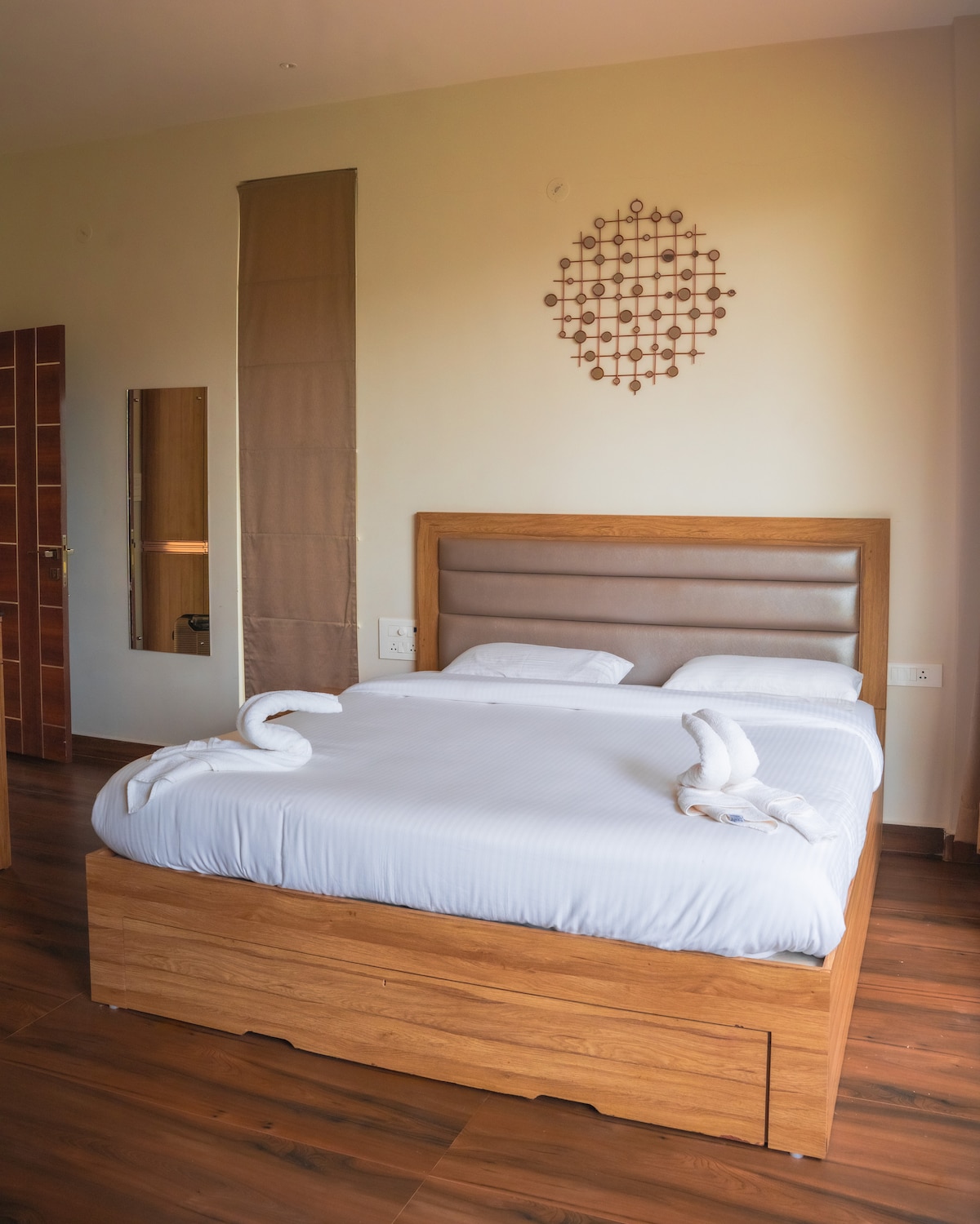 A spacious room features a wooden bed with a neutral-toned headboard, dressed in crisp white bedding. Decorative towel art is arranged on the bed. A mirror is mounted on the wall, reflecting the soft natural light from the nearby window.