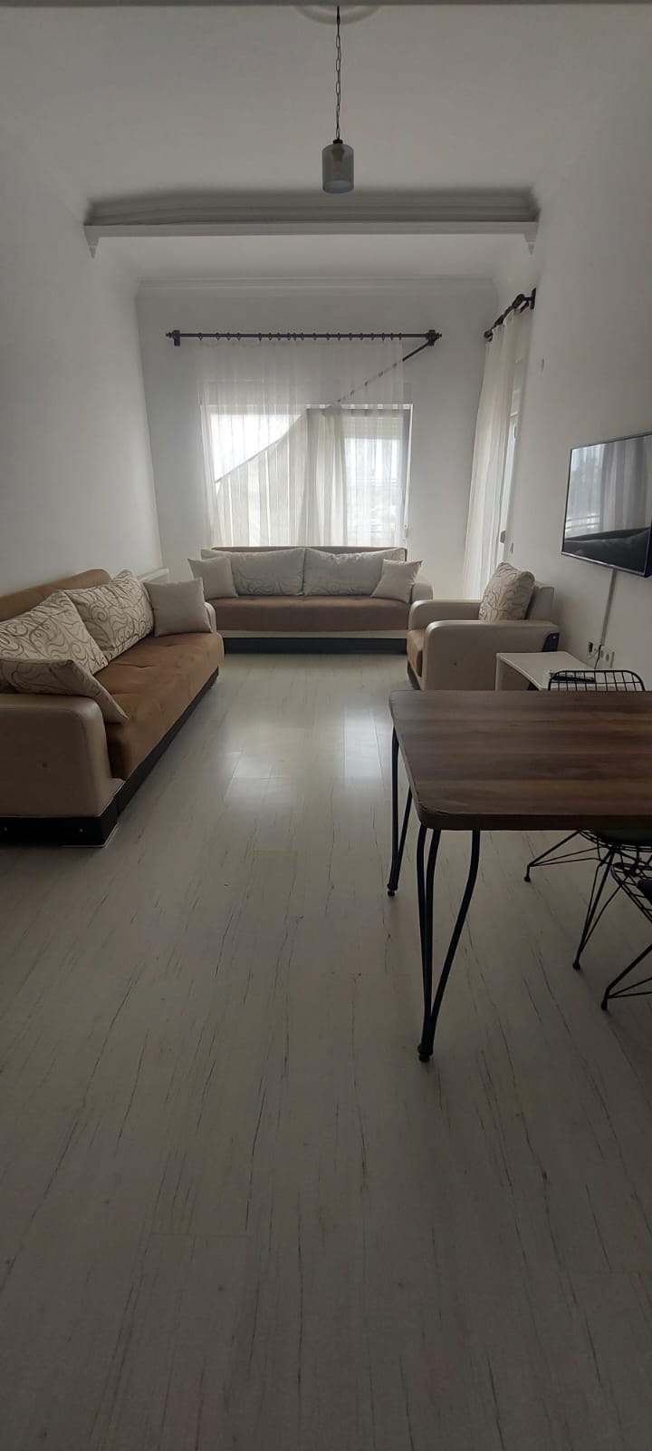 A light-filled living area features neutral-colored sofas arranged for comfort, accompanied by a small dining table with wire chairs. Sheer curtains allow natural light to flow in, enhancing the spacious feel of the room.