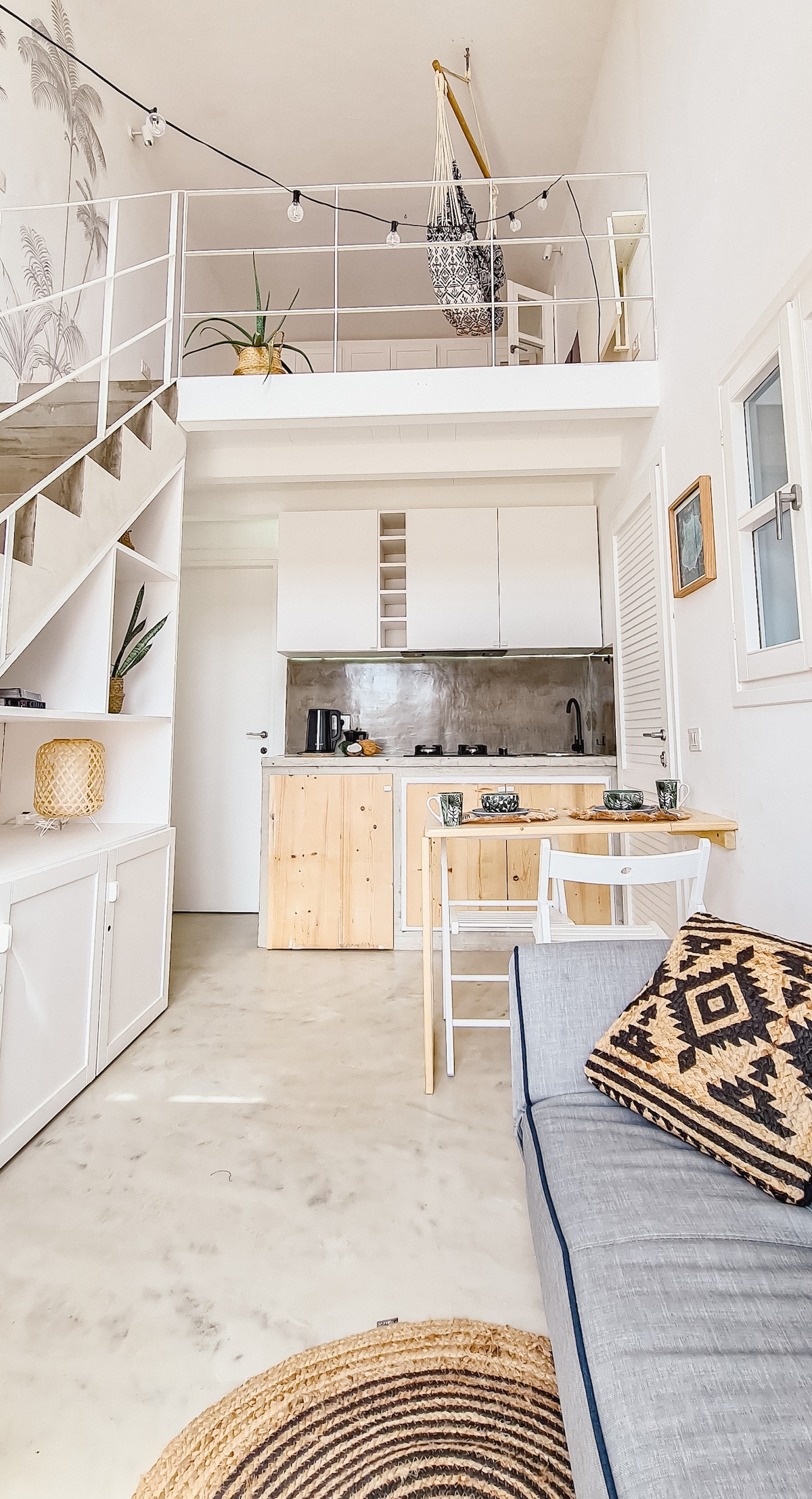The interior of the apartment displays an open-plan design with high ceilings. A well-equipped kitchenette is visible, paired with a dining table and chairs. The neutral tones are complemented by decorative elements like a woven rug and wall art, enhancing the light-filled atmosphere.