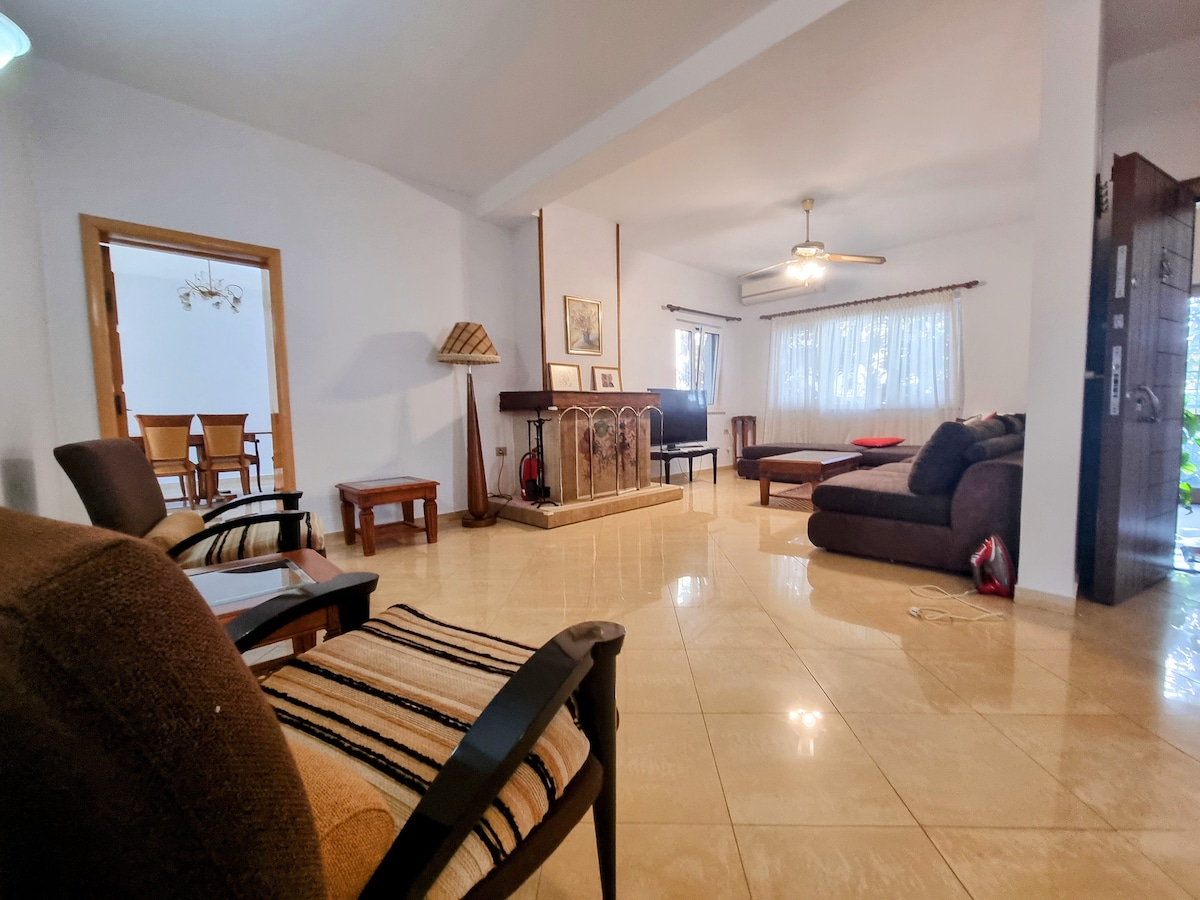 A spacious living area is highlighted by neutral-toned walls and tiled flooring. Comfortable seating options, including a dark sofa and patterned armchairs, are positioned near a decorative fireplace. Natural light filters through the window treatments, creating a bright and inviting atmosphere.