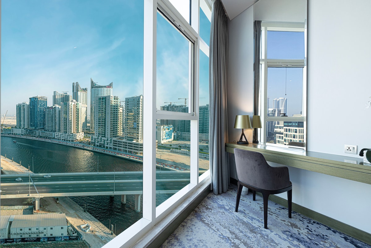 A modern workspace features a sleek desk and a stylish chair, set against large windows offering a panoramic view of a vibrant city skyline and canal. Natural light fills the space, highlighting the contemporary decor.