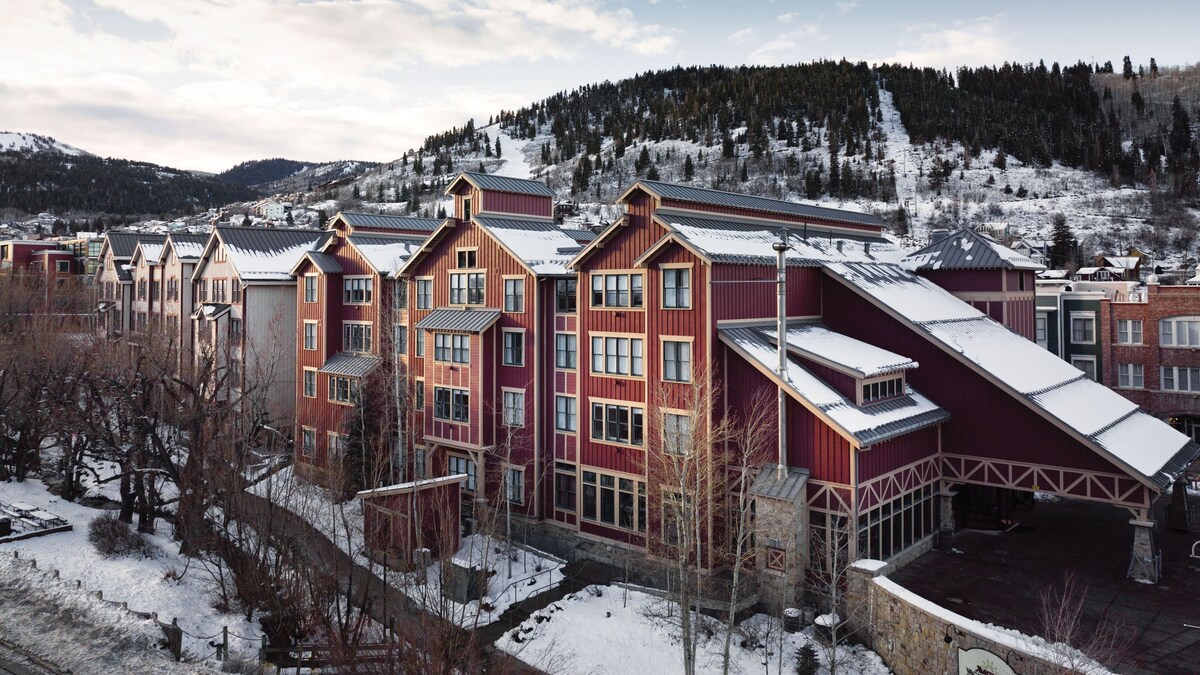 The exterior of a multi-story building is shown, featuring a combination of red and gray siding. Snow blankets the landscape, and tree branches create a natural border. The surrounding mountainous terrain is visible in the background, reflecting a serene winter atmosphere.
