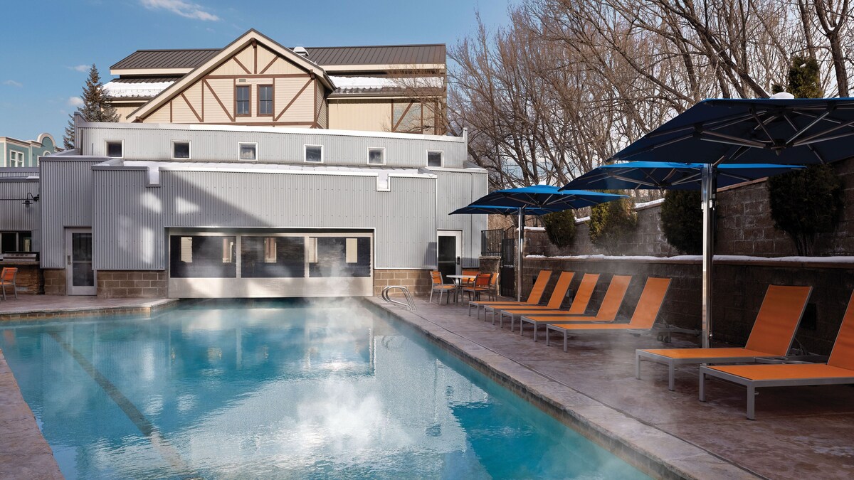 An outdoor heated pool is bordered by lounge chairs under blue umbrellas. Steam rises gently from the water, creating a serene atmosphere. A building with a distinctive design is visible in the background, complementing the peaceful setting.