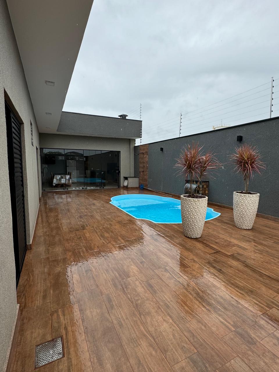 A spacious outdoor area features a private pool surrounded by textured tiles. Two large planters with ornamental plants are positioned near the pool. The covered space includes large glass doors, leading into the interior of the house, with a modern aesthetic emphasized by neutral tones.