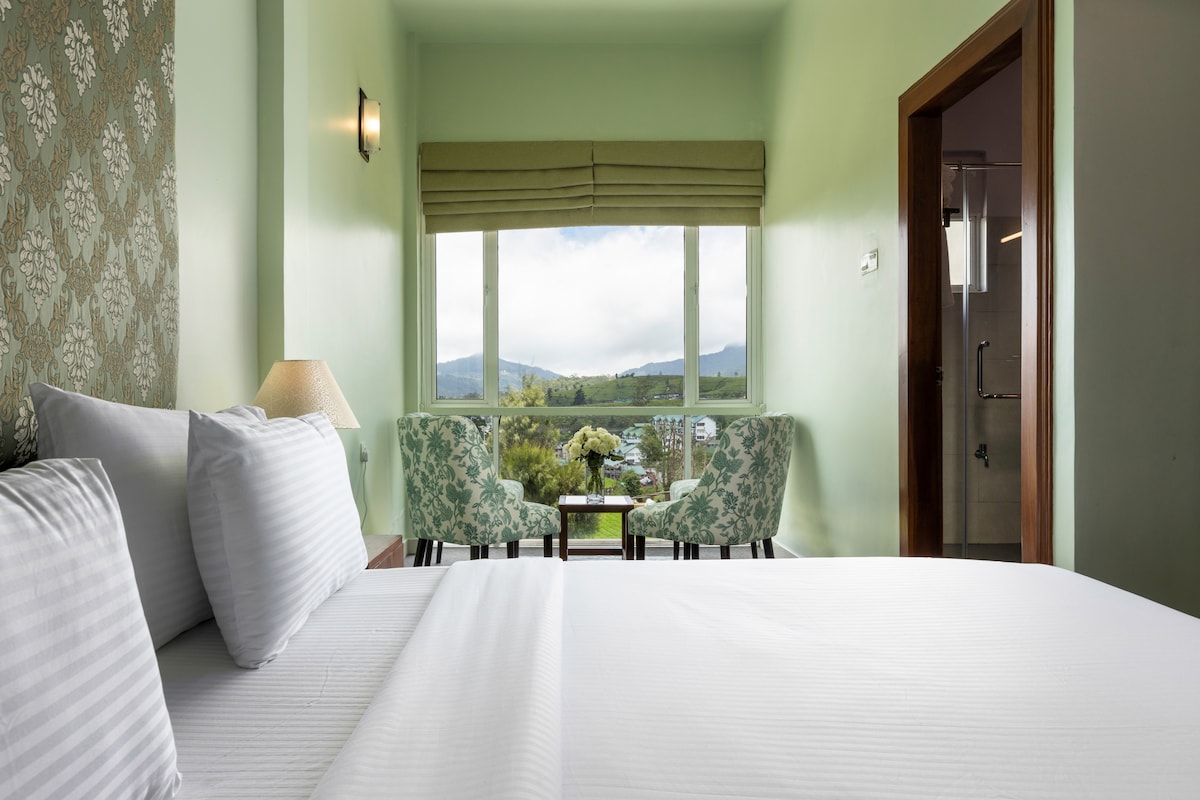 A spacious room features a neatly made bed with white linens, complemented by green patterned chairs flanking a small table near large windows. Soft natural light enters through the windows, highlighting the room's light green walls and enhancing the view of the surrounding hills.