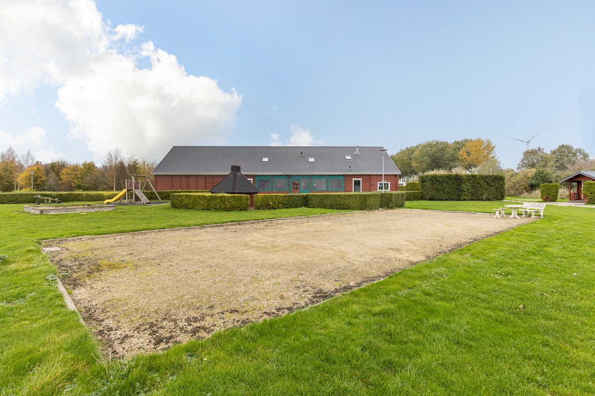 An outdoor area is presented with a well-maintained lawn and a large, level space suitable for activities. In the background, a chalet features multiple windows, while a playset with swings and a slide is to one side. Lush hedges provide natural boundaries.