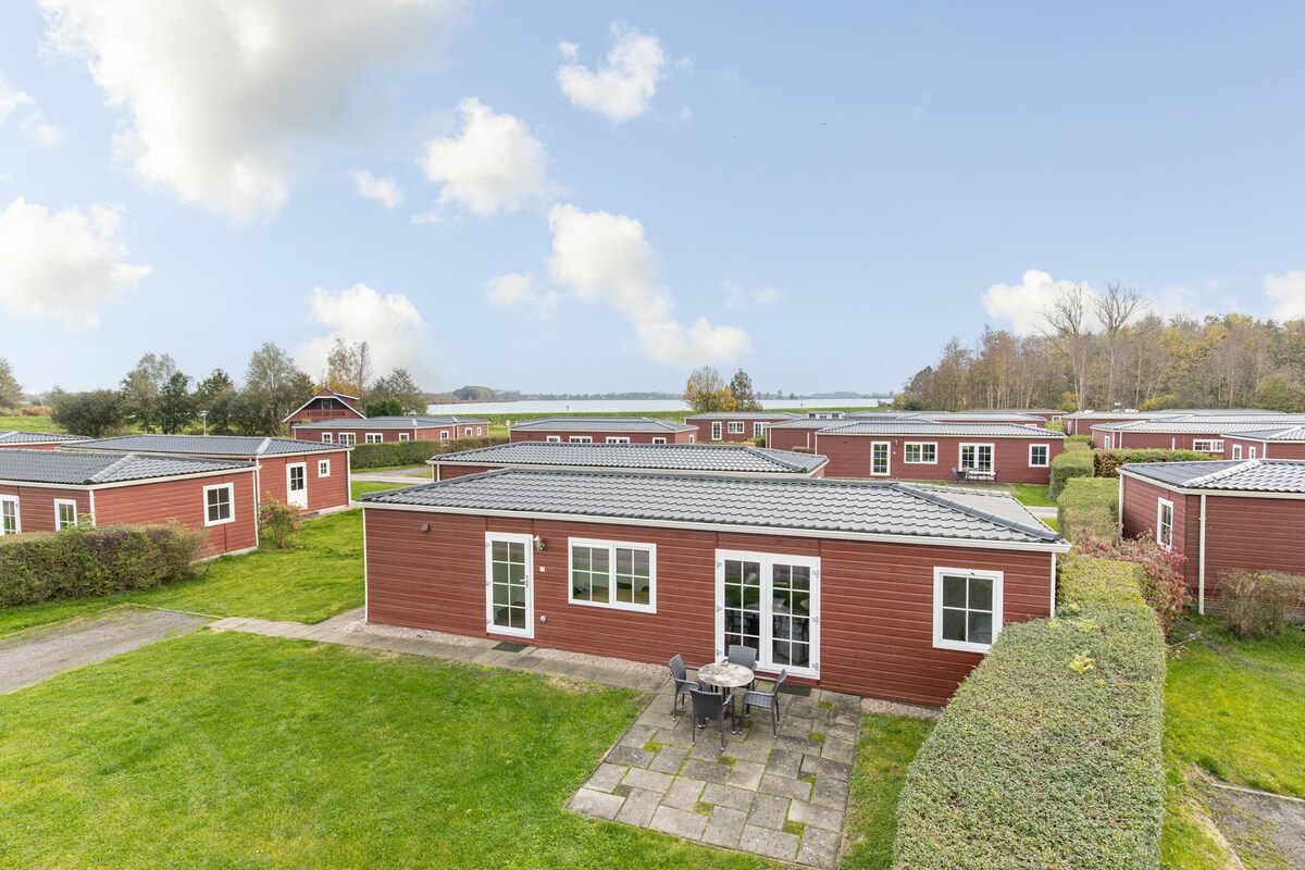 A view of multiple red chalets arranged around a green lawn, with a patio area featuring a small table and chairs in the foreground. The landscape is dotted with trees, and a body of water is visible in the background under a clear blue sky.