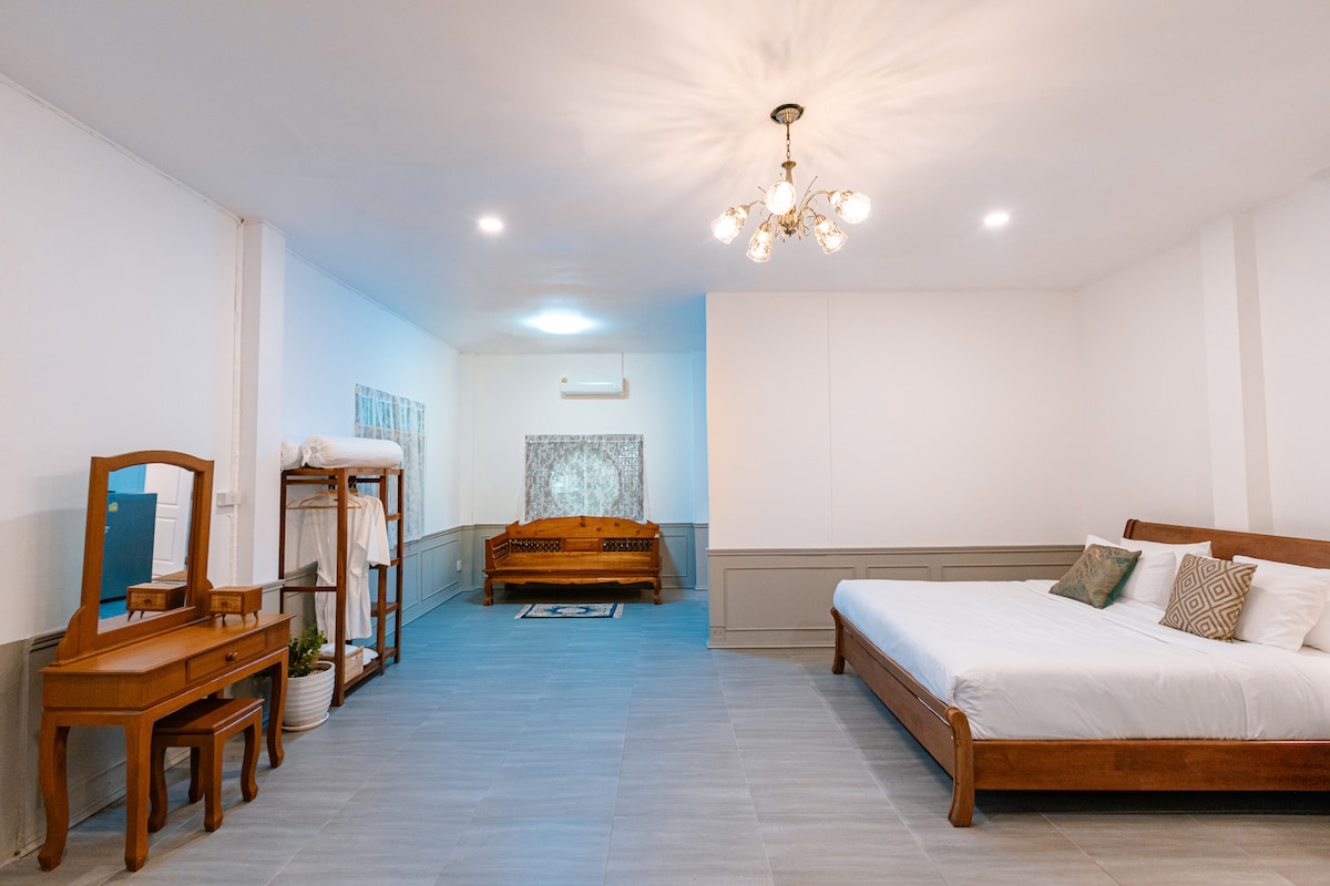 A spacious bedroom features a large bed with neatly arranged linens. A wooden vanity with a mirror is nearby, and a vintage seating area is situated in the corner. Soft lighting from a chandelier enhances the bright, airy atmosphere.