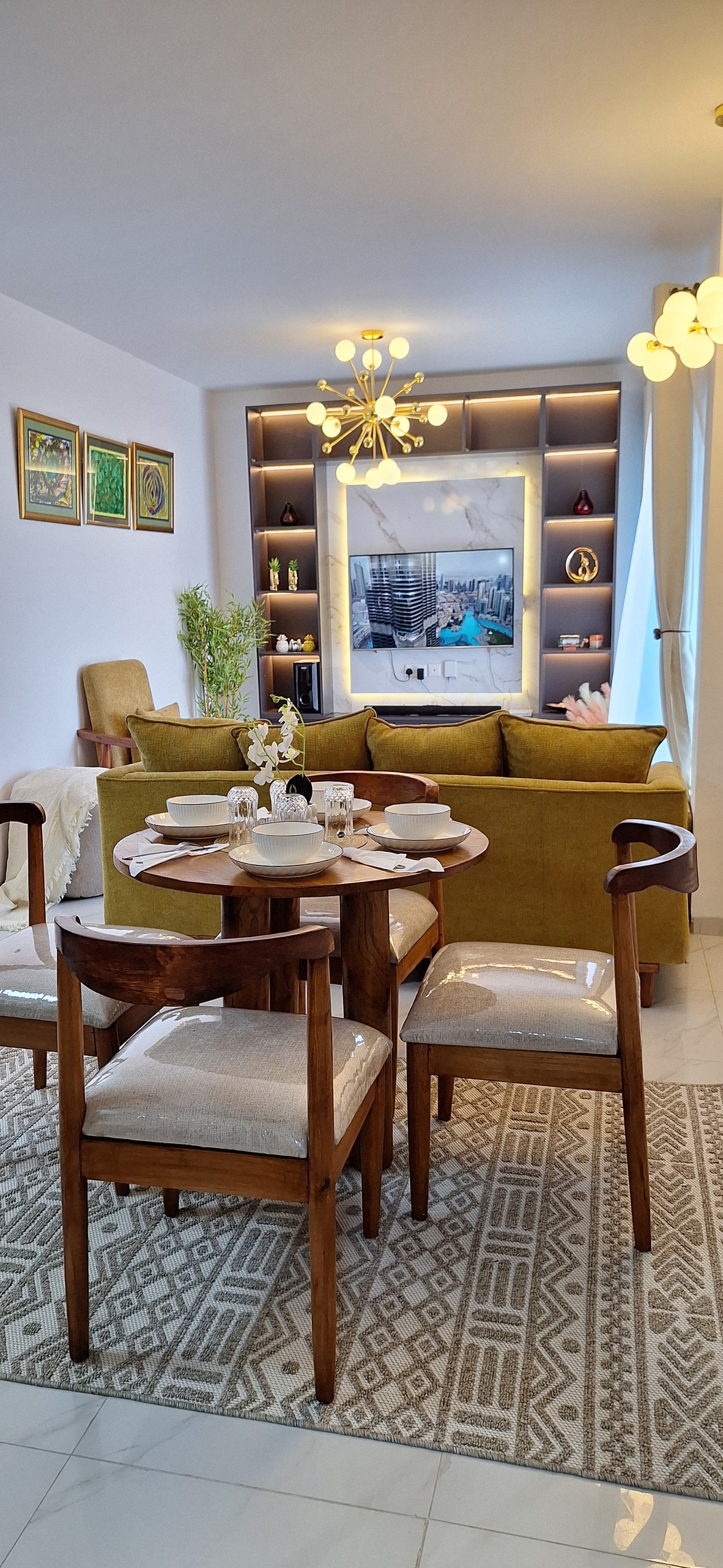 A cozy dining area features a wooden table set for four, surrounded by upholstered chairs. A soft rug lies beneath, and a green sofa is positioned nearby. A stylish light fixture hangs above, illuminating a framed television and decorative shelves in the background.