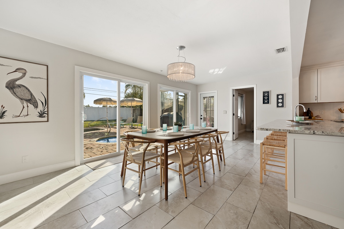 A bright dining area is furnished with a long wooden table surrounded by eight chairs. Natural light enters through large sliding glass doors, which provide a view of the backyard featuring a pool. A modern chandelier hangs overhead, complementing the open space.