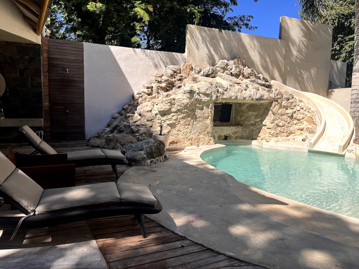 A private outdoor area features a curved swimming pool with a natural stone slide. Two lounge chairs are positioned beside the pool on a wooden deck, with lush greenery visible in the background, creating a serene and secluded environment.