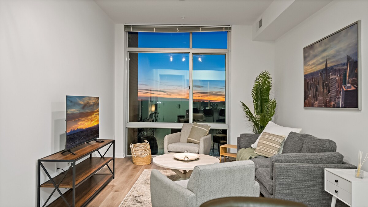 A modern living room features a seating area with two light grey chairs and a stylish sofa, complemented by a circular coffee table. Large windows offer city views, with a sunset reflecting vibrant colors. A television is positioned on a media console against a white wall.