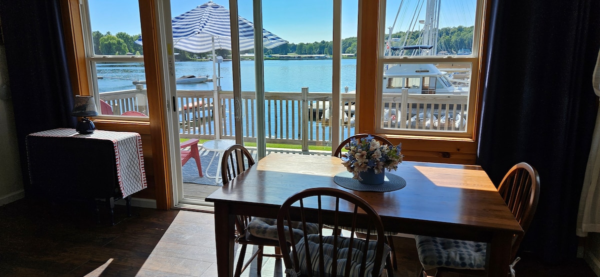 A dining area is visible, featuring a wooden table surrounded by several chairs. Large windows offer a view of the water and boats outside, with a striped umbrella casting shade over the deck. A small floral arrangement adorns the center of the table.