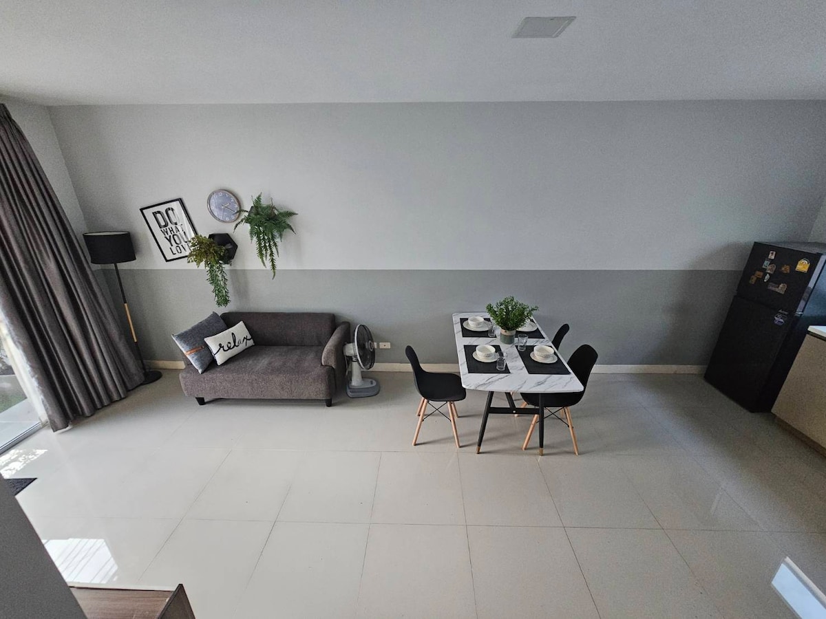 An open living and dining area is visible, featuring a modern sofa and a dining table set for four. Greenery decorates the space, complemented by sleek, minimal furnishings. Large windows allow natural light to illuminate the polished flooring, enhancing the airy feel of the room.