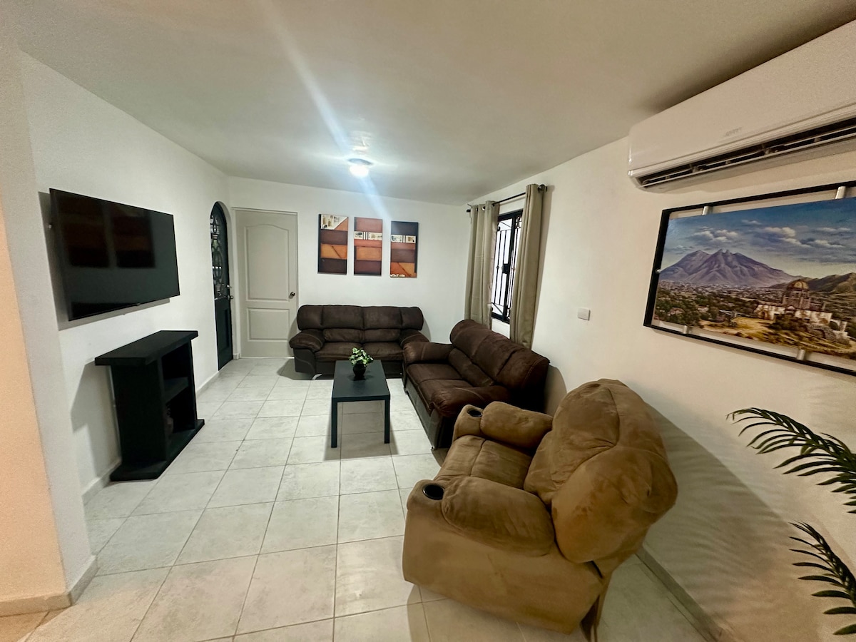 A comfortable living area is presented with a sectional sofa, two plush armchairs, and a coffee table. A wall-mounted TV is positioned on one side, while a decorative painting hangs above the furniture. Large windows allow natural light to brighten the white walls and tiled floor.