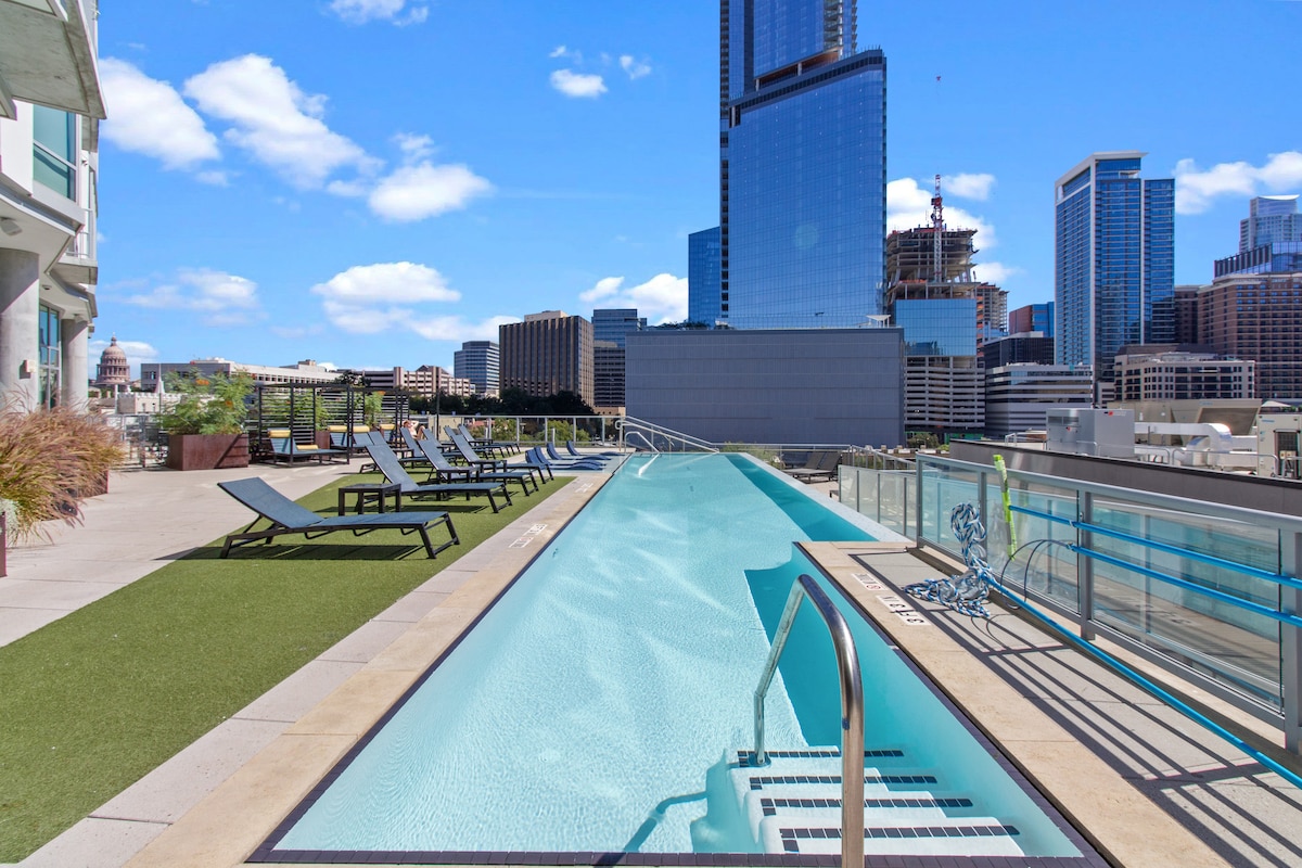 A rooftop pool area is visible, featuring a long, modern pool with gentle steps leading in. Surrounding loungers are positioned on a green turf area, with a city skyline providing a vibrant backdrop against a clear blue sky.