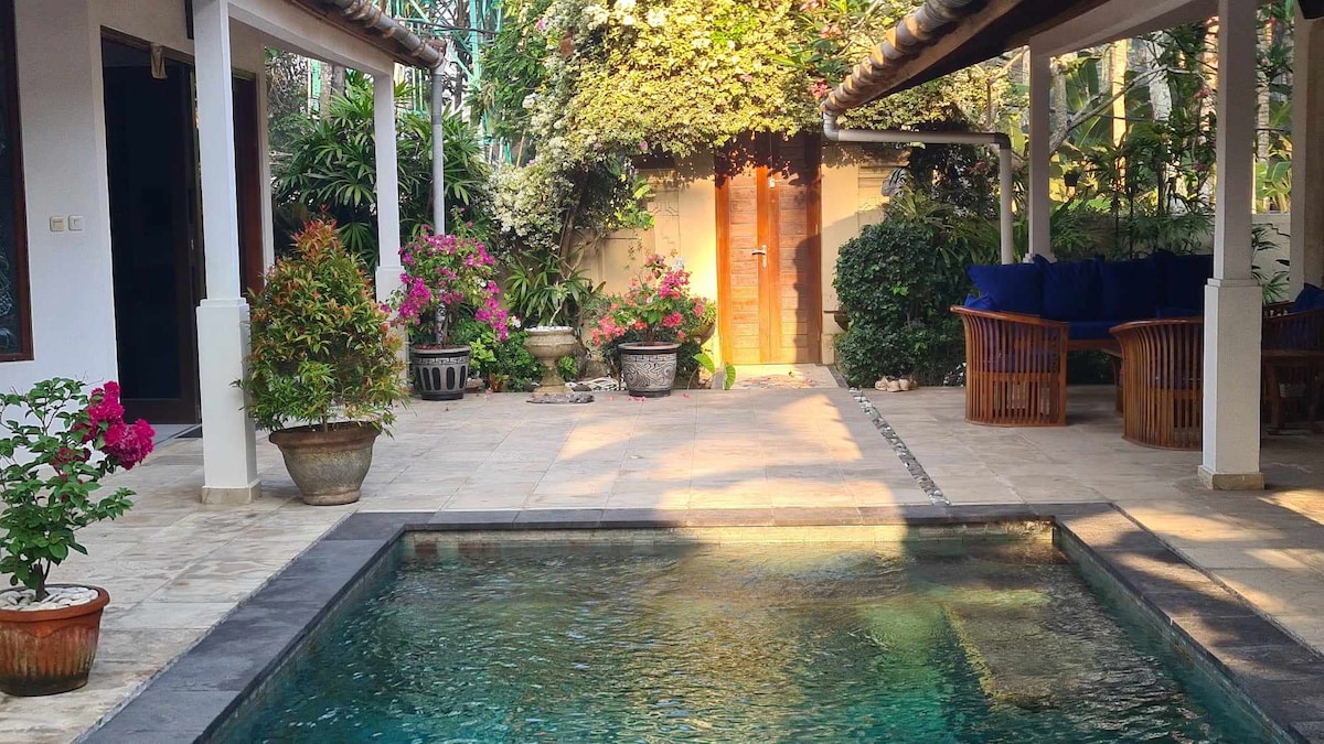 The image presents a serene outdoor area featuring a swimming pool surrounded by stone tiles and lush greenery. Vibrant flowering plants in pots add color, while a covered lounging space with comfortable seating is visible. A wooden door in the background suggests access to another area.