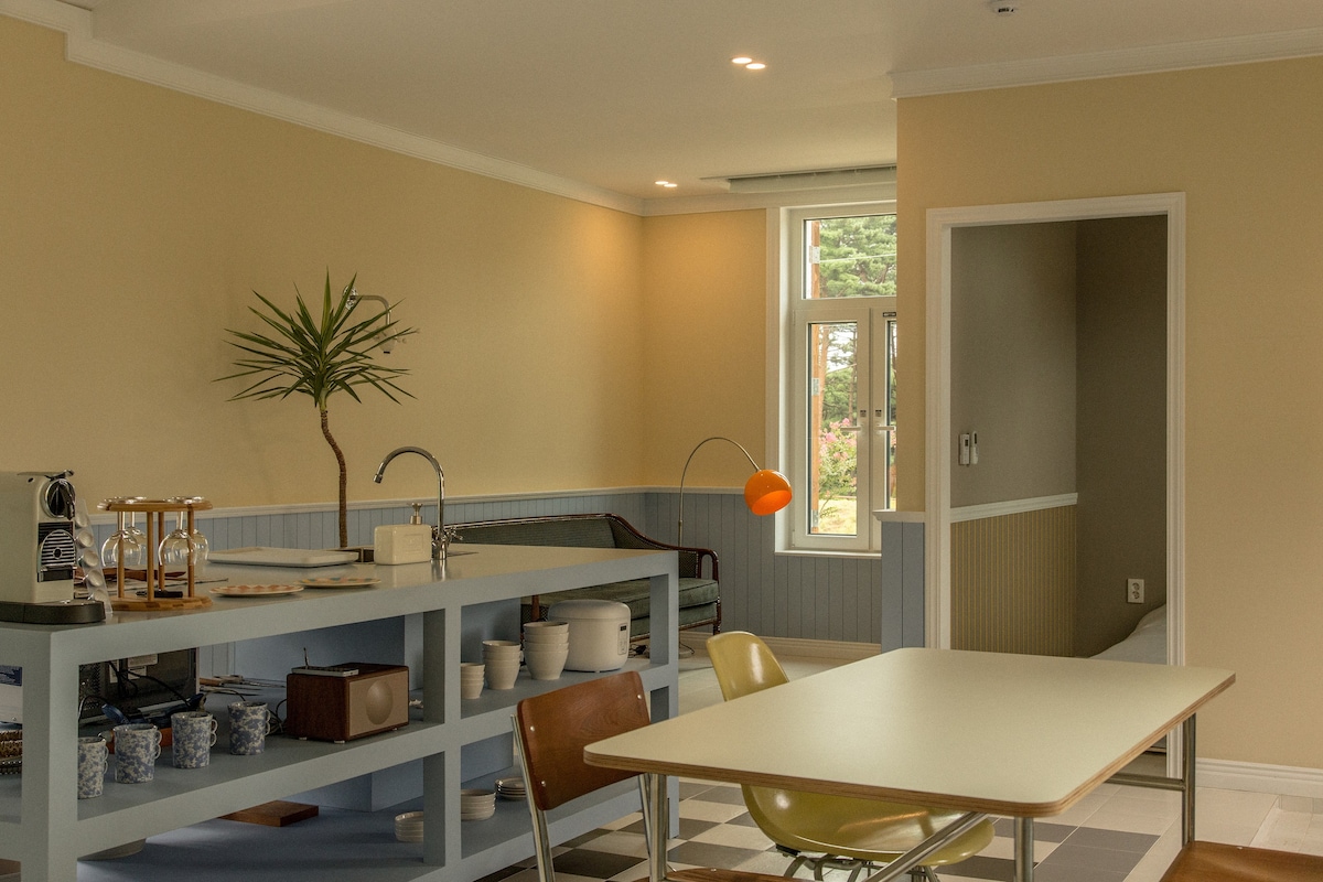 An open kitchen area features light pastel tones, complemented by a modern table and chairs. A plant adds a touch of greenery, while large windows allow natural light to fill the space. A cozy seating nook is visible in the background.