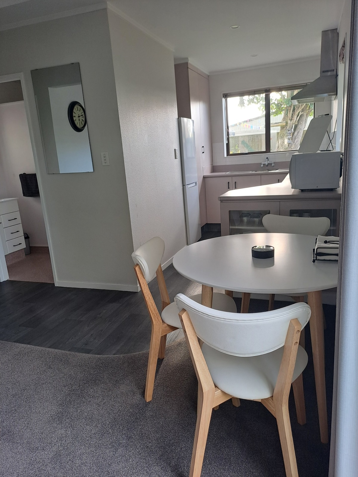 A dining area is featured with a round table and three chairs, set against a neutral backdrop. A window provides natural light, overlooking the exterior trees. Kitchen appliances are visible in the background, with cabinetry and a mirror enhancing the space's functionality.