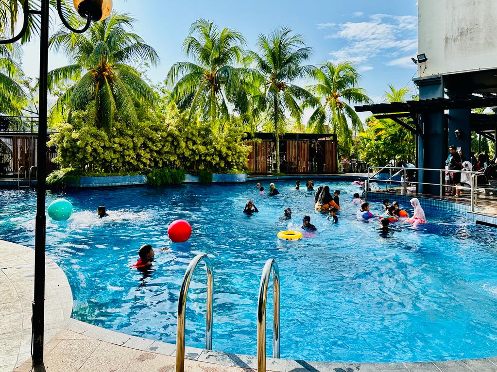 A vibrant swimming pool scene is depicted, filled with people enjoying the water. Lush palm trees surround the area, creating a tropical atmosphere. Colorful floatation devices can be seen, adding to the recreational vibe. Sunlight dapples the water’s surface, enhancing the inviting environment.