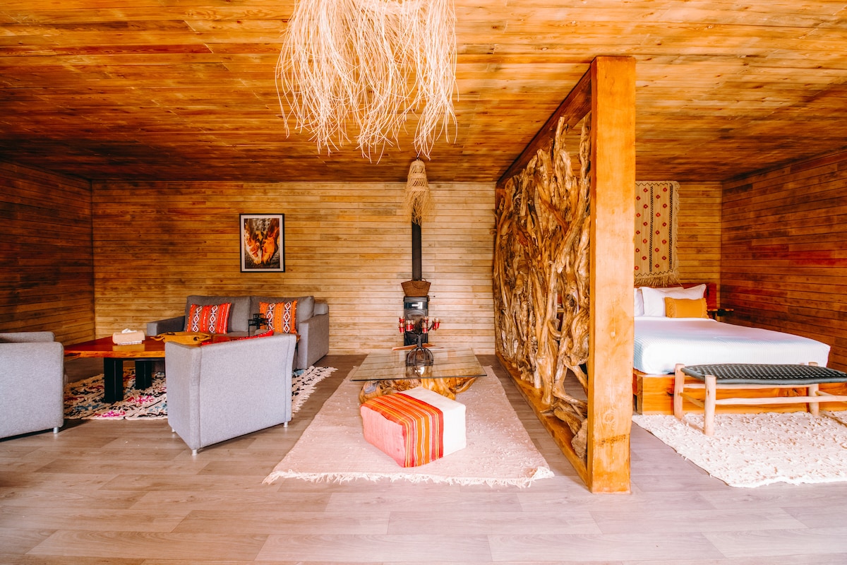A spacious room is characterized by wooden walls and a warm, natural design. The area features a comfortable bed, a cozy seating arrangement with soft gray sofas, and a unique wooden art piece dividing the space. A rustic wood stove and decorative rugs enhance the ambiance.