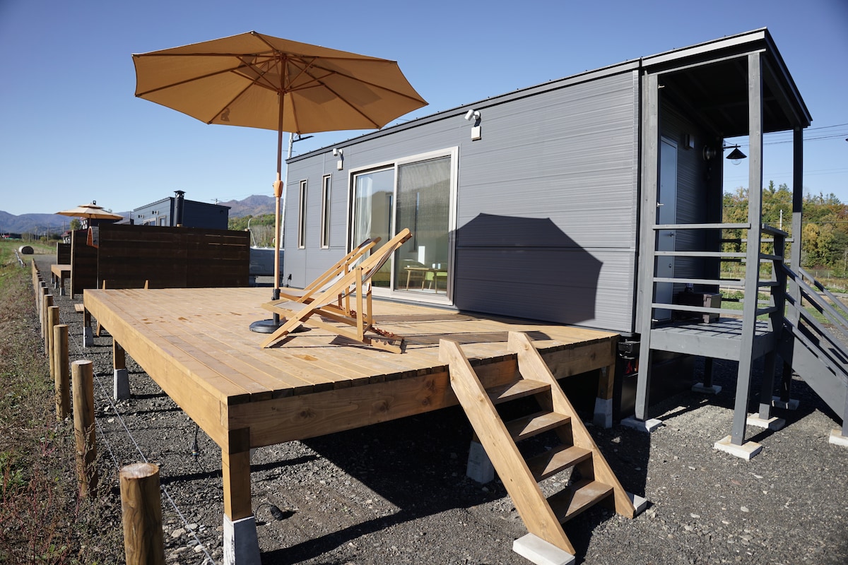 A spacious wooden deck is featured outside the trailer house, equipped with a large parasol that provides shade. A lounge chair is positioned on the deck, with steps leading up to the entrance of the accommodation. Surrounding scenery includes distant mountains and clear skies.