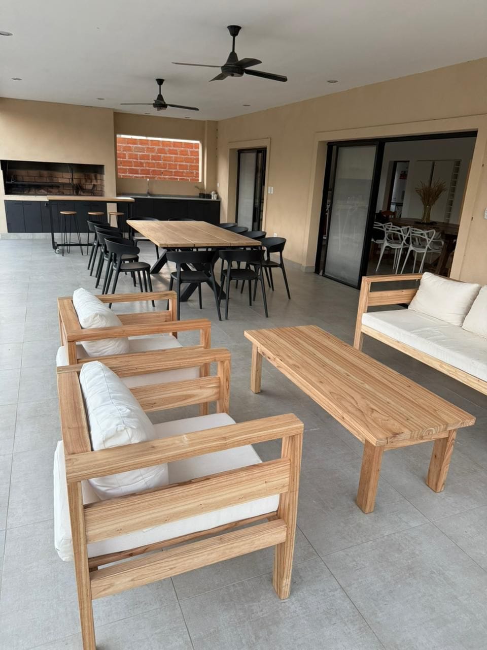 An open-air terrace features a blend of wooden and black dining furniture. A spacious table is surrounded by black chairs, while a comfortable seating area includes light-colored couches and a wooden coffee table. Ceiling fans are positioned above for ventilation.