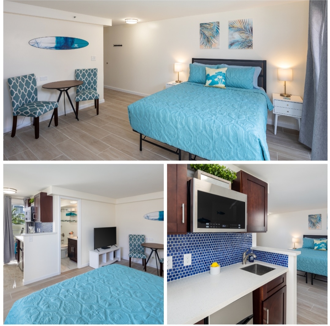 A spacious corner studio is featured, showcasing a memory foam queen bed layered in a soothing blue coverlet. A small table with two chairs occupies one corner, while a kitchenette is visible, equipped with modern appliances and tiled backsplash.