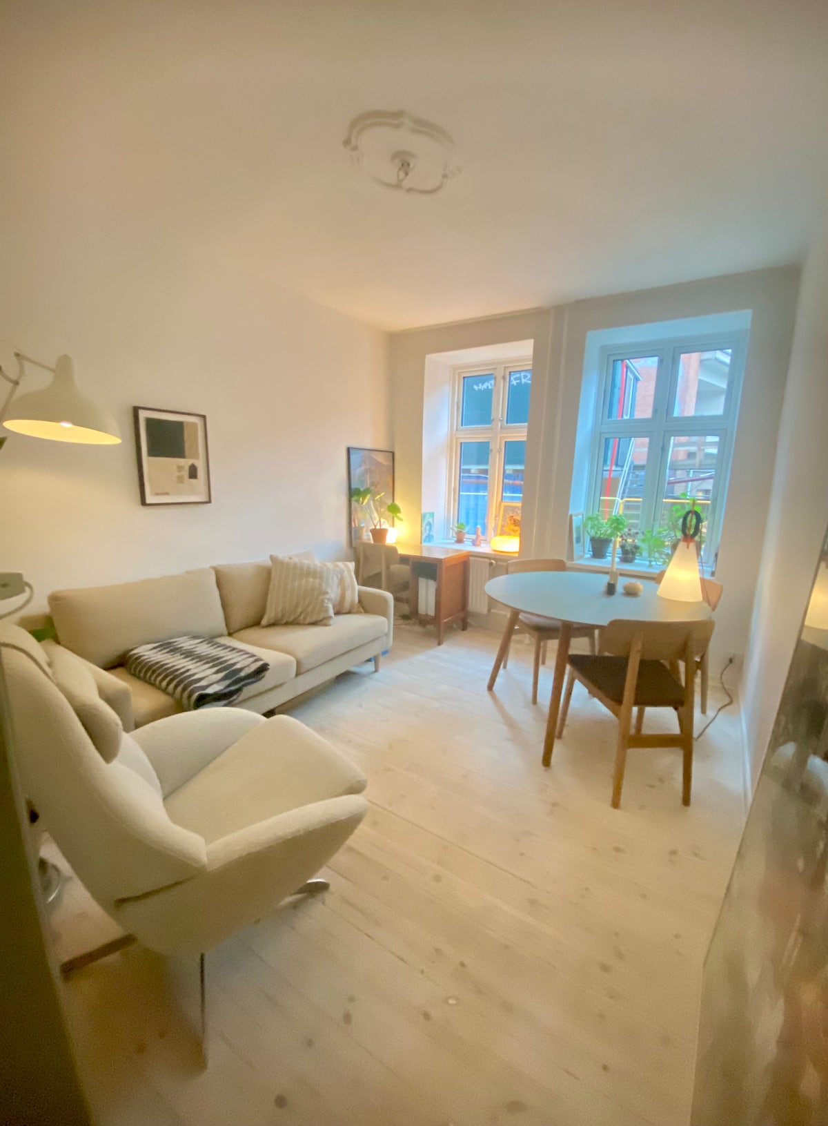 A light-filled living area is characterized by two large windows draped with light curtains. A cozy sofa is accompanied by a textured throw and cushion. A round dining table surrounded by wooden chairs and a lamp creates a warm, inviting space for relaxation or work.