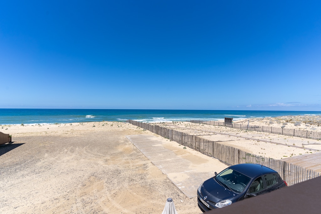 A panoramic view of the ocean is captured, showcasing a clear blue sky and gentle waves lapping at the shore. The sandy beach stretches into the distance, bordered by a wooden fence, with a parked car visible in the foreground.