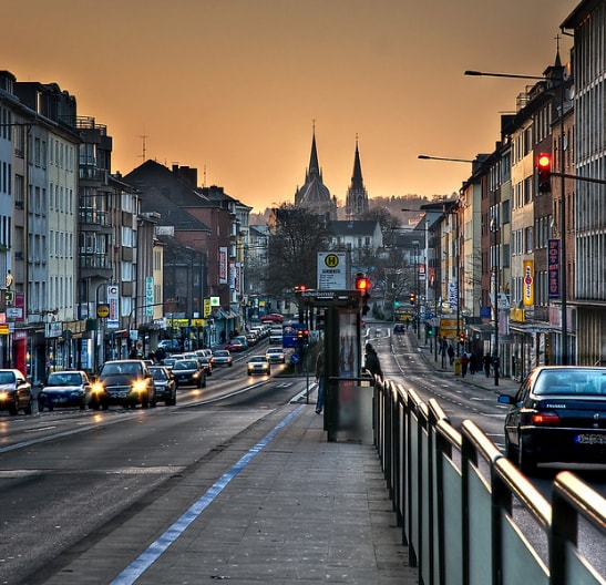 A busy street scene showcases a well-maintained roadway lined with various buildings. The silhouette of two church spires is visible against the sunset, adding depth to the urban landscape. Streetlights illuminate the area, while vehicles are seen traveling along the thoroughfare.