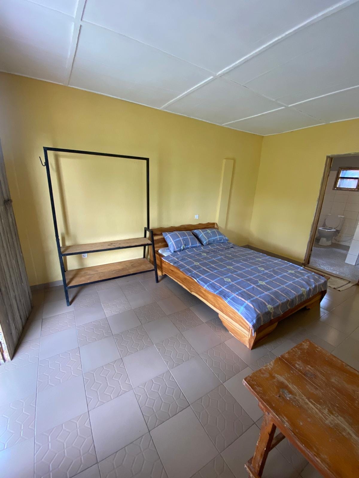 A simple bedroom features a wooden bed with a blue patterned blanket. A clothes rack stands nearby, and a wooden table is positioned in the corner. Soft natural light illuminates the space, highlighting the neutral-toned walls and tiled floor.