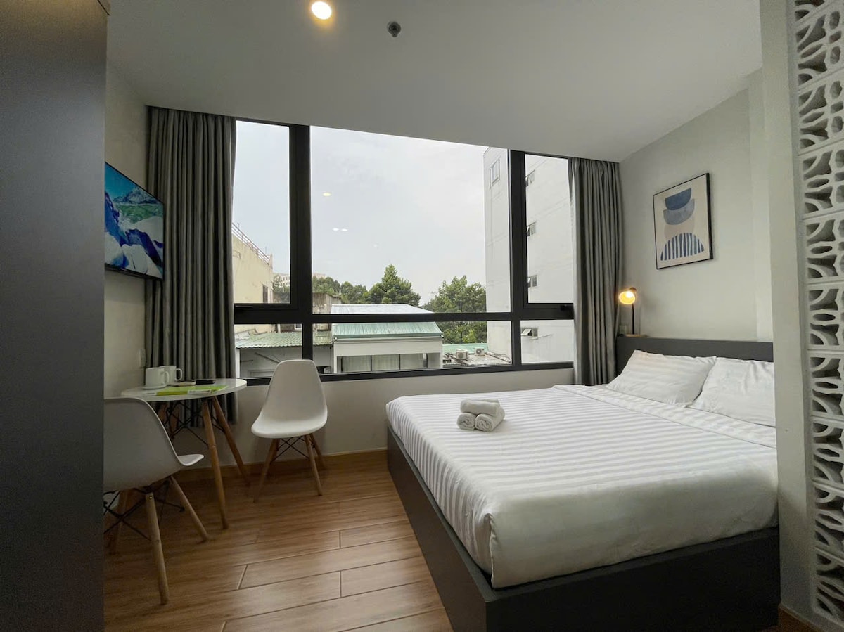A modern studio room features large windows allowing natural light to enter, framed by neutral-toned curtains. A spacious bed is showcased alongside a small table with two chairs. A smart TV is mounted on the wall, adding functionality to the comfortable space.