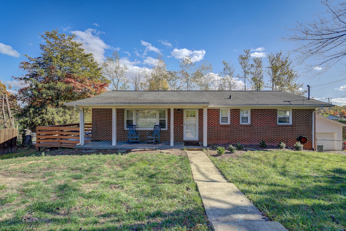 The exterior of the home is showcased with a brick facade and a covered front porch featuring two blue chairs. A concrete path leads to the entrance, framed by a well-maintained lawn with greenery and landscaping. Trees in the background provide a natural setting.