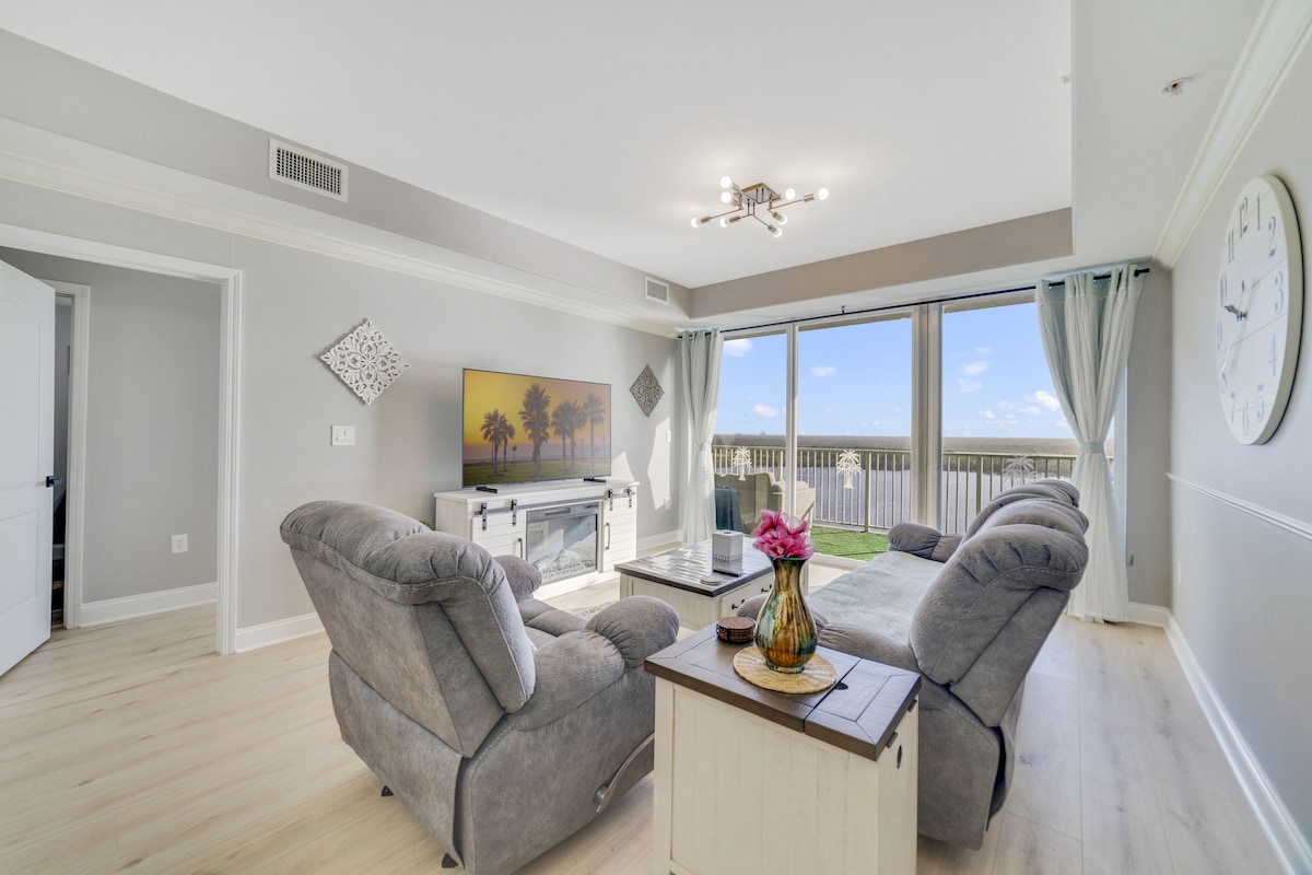 A spacious living area features comfortable grey recliners and a light wooden coffee table. A large screen TV is mounted above an entertainment console, while expansive windows allow natural light to fill the room. Views of the balcony and outdoors can be seen through the sheer curtains.